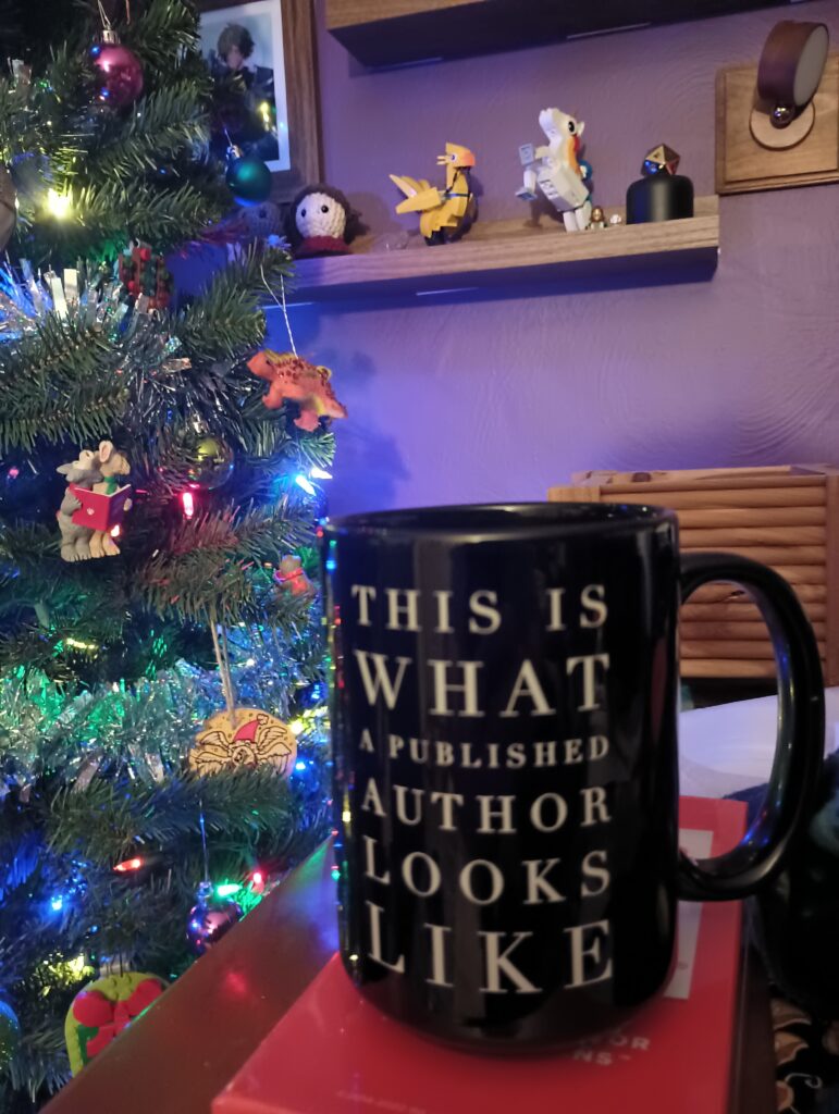 A coffee mug in front of a Christmas tree.  The text on the mug reads "This is what a published author looks like"