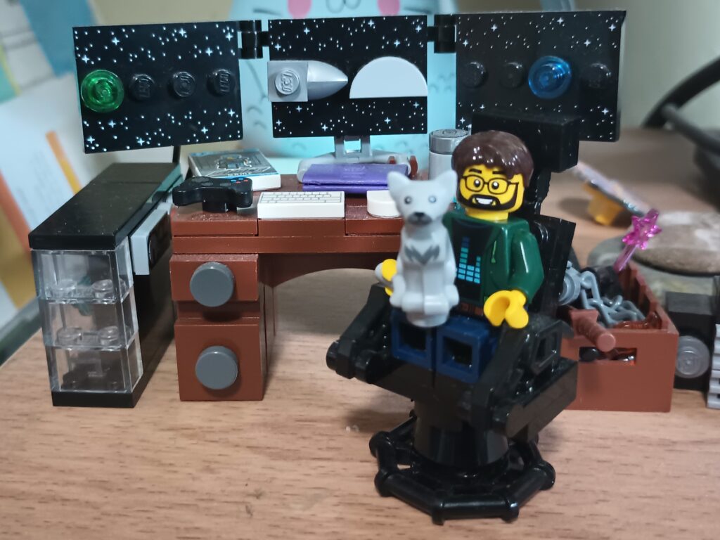 A Lego Minifigure sitting at a computer desk with a Lego Cat in their lap.